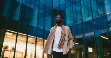 Tarz sahibi Afrikalı Amerikalı adam modern yüksek binadan rahatça uzaklaşıyor. Arka plandan geçen bir kadın. İşten sonra planlama akşamı. Soğuk hava. Gözlük takıyordu. Etrafa bakınıyorum..