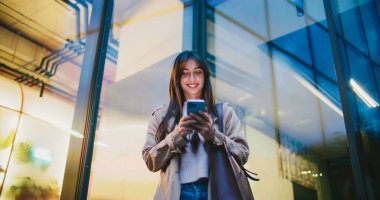 Çekici beyaz kadın akşamları modern ticaret merkezinden ayrılıyor. Akıllı telefonunu kullanıyordu. Ceket ve kazak giyiyor. Yeni şehirde turist olarak pozitif bir akşam. İlginç konumlar araştırılıyor.