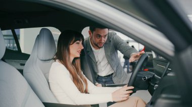 Güzel, beyaz bir kadın, satış müdürüyle konuşurken arabada oturuyor. Sorular soruyor. Cevap vermeden önce tablet cihazına bakan adam. Araç ekranındaki düğmeye bir kez basılıyor.