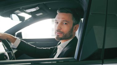 Takım elbiseli yakışıklı beyaz bir erkek arabada oturuyor ve yeni teknolojiye hayran. Sürücü aracını yolda hayal ediyorum. Yerel bayiyi ziyaret ediyorum. Doğrudan bakıp gülümsüyor.