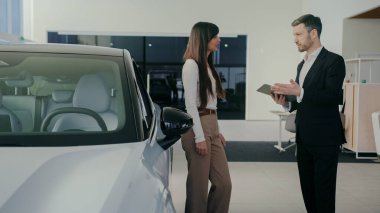 Güzel beyaz kadın araba galerisinde bulunan yeni SUV 'ye ilgi gösteriyor. Resmi kıyafet mağazasının müdürü kapıyı açıyor ve aracın içini gösteriyor. Dinlerken içeri bakan kadın..