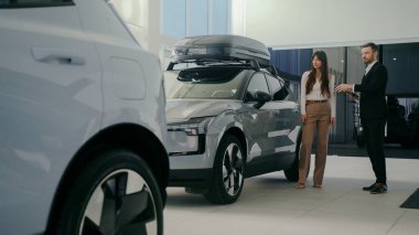 Modern arabanın yanında durup müdürün konuşmasını dinleyen sevimli beyaz bir çift. Sakallı erkek, yeni modelle ilgili tüm detayları açıklıyor. Çatıdaki ek depoyu parmakla göstermek.