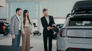 Profesyonel satış müdürü müşterilerinin ilk elektrikli aracını seçmesine yardım ediyor. Aracın yan tarafındaki şarj kapağı açılıyor ve işlem açıklanıyor. Soruları cevaplıyorum. İnsanlara yardım etmek.