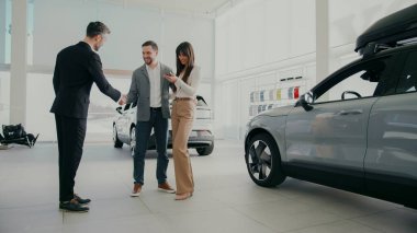 Araba galerisinde kadın ve erkek yeni arabanın yanında durup satıcıyla alışverişlerini tartışıyorlar. Torbacı arabasının anahtarlarını verirken elini uzatan kadın. İnsanlar seçimlerinden memnun..