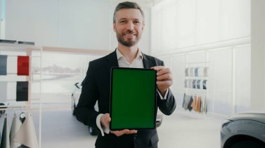 Resmi takım elbiseli yakışıklı adam oto kuaförde duruyor ve gülümsüyor. Yeşil ekranlı tablet cihazı tutan mutlu işçi. Arka planda görünür araba ve kumaş örnekleriyle dolu raflar var. Çalışma ortamı.