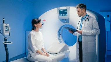Dikkatli esmer kadın MRI tarayıcısının yanında oturuyor ve doktorların sorularına cevap veriyor. Ciddi bir tıbbi uzman pano tutuyor ve tanılama prosedürüne hazırlanırken dikkatlice dinliyor..