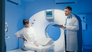 Beyaz kadın elini sırtına koyuyor ve muayene sırasında acı belirtilerini tarif ediyor. Görüntüleme ekipmanlarının yanında dururken panoya gözlemler yazan odaklanmış erkek doktor.