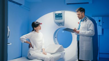 Ciddi bir beyaz kadın tomografide oturuyor ve odaklanmış erkek doktorun beyin taraması sonuçlarını dikkatlice dinliyor. Tıbbi görüntülere bakmak ve muayene sırasında teşhisi açıklamak..