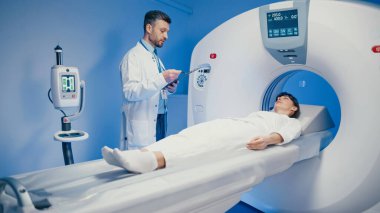 Kafkasyalı rahat bir kadın tomografi çekilirken tomografi masasında uzanıyor ve dikkatli bir erkek doktor notları inceliyor. Hastane görüntüleme odasındaki radyoloji oturumunu denetlerken kalem ve pano tutuluyor.