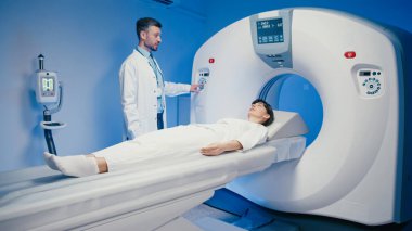 Beyaz erkek doktor tomografi cihazını çalıştırırken tarama detaylarını kadınlara açıklıyor. Kadın hasta muayene masasında, gözleri kapalı, tıbbi görüntüleme işlemine hazır yatıyor..