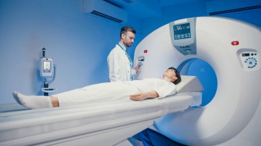 Kafkas kadın CT tarama yatağında hareketsiz yatıyor. Tanısal görüntüleme için makineye kayıyor. Odaklanmış erkek teknisyen muayene odasında tıbbi ceket giyerken düğmelere basıyor ve prosedürü izliyor.