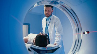 Yakışıklı erkek doktor tomografi tünelinde hastaya bir şey açıklarken elini kaldırıyor. Prosedür sona ererken tarama masasında yatan beyaz kadın. Makinede parlayan lazer hattı.