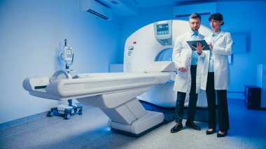 Çekici kadın doktor elinde tabletle meslektaşlarına sonuçları açıklıyor. Raporu incelerken CT makinesinin yanında dikkatle dinleyen beyaz erkek uzman. Radyoloji odasında birlikte duruyoruz..