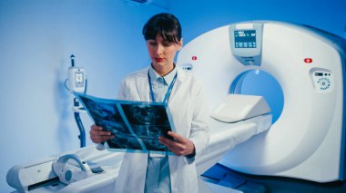 Ciddi bir beyaz kadın teknisyen radyoloji odasında MRI tarayıcısının önünde duruyor. Teşhis prosedürüne ya da bir sonraki hastaya hazırlanıyorum. Çok ciddi bir ifadeyle doğrudan kameraya bakıyor..