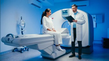 Tarama masasında oturan huzurlu kadın laboratuvar önlüklü erkek doktoru dinlerken başını sallıyor. MRI ünitesi yakınlarındaki tıbbi brifing sırasında, prosedür veya teşhisi açıklayan profesyonel bir dosya..