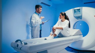 Gülümseyen erkek doktor, MRI masasında oturan neşeli bir hastayla dostça sohbet ediyor. Pano tutuluyor ve teşhis odasındaki tıbbi taramadan önce güvence veriliyor..