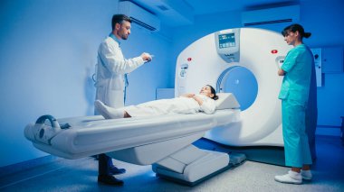 Kendine güvenen erkek doktor panoya yazı yazarken yakından gözlemliyor. Üniformalı kadın teknisyen, hasta yaklaşan muayene için yavaşça tarayıcıya doğru ilerlerken yatak kaldırma işlevini etkinleştiriyor..