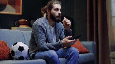 Kanepede minderlerle oturan bir erkek. Sakallı adam elinde telefonla televizyon izliyor. Erkek futbol maçı izliyor. Favori takım için neşelenen bir adam. Futbol topu şeklinde yastık..