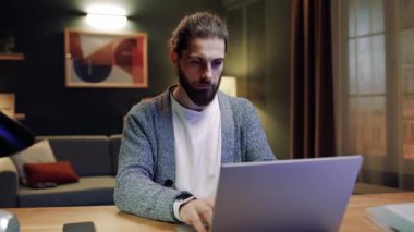 Oturma odasında masada oturan bir erkek. Evde dizüstü bilgisayarla çalışan bir adam. Erkek çevrimiçi çalışıyor. Serbest çalışmak için dizüstü bilgisayar kullanan bir adam. Erkek kameraya bakıyor. Akıllı saat takan adam klavyede yazı yazıyor..