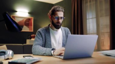 Oturma odasında masada oturan bir erkek. Evde dizüstü bilgisayarla çalışan bir iş adamı. Uzaktan çalışan bir erkek. Serbest çalışmak için dizüstü bilgisayar kullanan bir adam. Gözlüklü bir erkek kameraya bakıyor. Klavyede yazan adam.