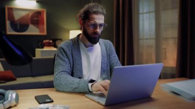 Oturma odasında masada oturan bir erkek. Evde dizüstü bilgisayarla çalışan bir iş adamı. Uzaktan çalışan bir erkek. Serbest çalışmak için dizüstü bilgisayar kullanan bir adam. Gözlüklü, konsantre bir erkek. Klavyede yazan adam.