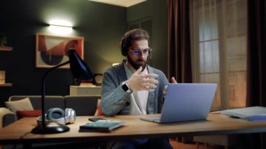 Oturma odasında masada oturan bir erkek. Çevrimiçi toplantı yapan bir adam. Uzaktan çalışan bir erkek. Video görüşmesi için laptop kullanan bir adam. Erkek, gözlük ve kulaklık takıyor. Klavyede yazan adam.