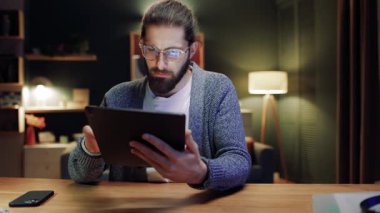 Oturma odasında elinde tablet cihazıyla oturan gözlüklü çekici bir adam. Telekomünikasyon işi, sosyal medyada sohbet, web sitelerinde arama. E-ticaret, sanal hizmetler, eğlenceli uygulama kavramı