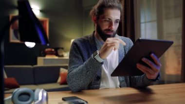 Genç yakışıklı adam akşam evde oturma odasında otururken modern tablette video akışı izliyor. Gülümseyen erkek çevrimiçi yayın izliyor ya da bilgisayar oyunu oynuyor.