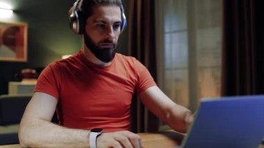 Oturma odasında masada oturan bir erkek. Evde dizüstü bilgisayarla çalışan bir iş adamı. Uzaktan çalışan düşünceli bir erkek. Adam iş için dizüstü bilgisayar açıyor. Kulaklıklı, odaklanmış bir erkek. Klavyede yazan adam.