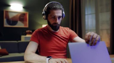 Oturma odasında masada oturan bir erkek. Evde dizüstü bilgisayarla çalışan bir iş adamı. Uzaktan çalışan düşünceli bir erkek. Adam dizüstü bilgisayarı kullanmak için açıyor. Kulaklıklı, odaklanmış bir erkek. Evden çalışan adam..