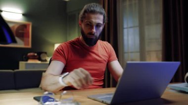 Oturma odasında masada oturan bir erkek. İş adamı yeni projeler yaratıyor. Uzaktan çalışan odaklanmış bir erkek. Serbest çalışmak için dizüstü bilgisayar kullanan bir adam. Ciddi bir adam laptop ekranına bakıyor. Gözlük takan adam.