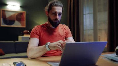 Oturma odasında masada oturan bir erkek. İş adamı yeni projeler yaratıyor. Uzaktan çalışan bir erkek. Serbest çalışmak için dizüstü bilgisayar kullanan bir adam. Ciddi bir adam laptop ekranına bakıyor. Klavyede yazan adam.