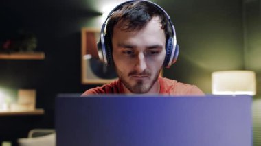 Genç adam dizüstü bilgisayarın önünde oturuyor. Laptop ekranına bakan tatmin olmuş bir adam. Erkek evde dinleniyor. Oyun oynamak için dizüstü bilgisayar kullanan gülümseyen adam. Kulaklık takan ve klavye çalan genç adam..