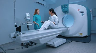 Radyologun bilgisayarlı tomografi odasında hastası var. Nörolog doktor hastanın yanında duruyor ve prosedürü açıklıyor. Sağlık çalışanı konuşuyor, hasta doktoru dikkatle dinliyor..