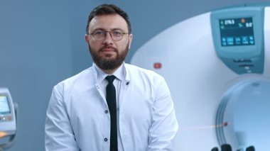 Tomografi kapsülünde oturan deneyimli bir doktorun portresi. Gözlüklü, sakallı bir doktor. MRI taraması için modern donanımlı oda. MRI kapsülünün arka planında tomograf.