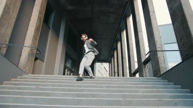 Takım elbiseli beyaz bir çalışan kulaklıkla müzik dinliyor, elinde akıllı telefon, merdivenlerden inerken dans ediyor. Mutluluk kavramı, iyi ruh hali, başarı