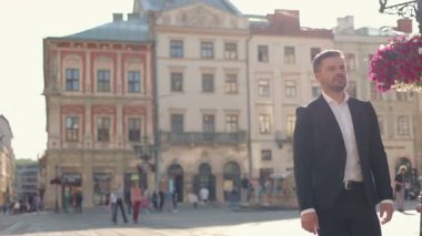 Beyaz, Avrupalı, orta yaşlı, takım elbiseli bir işadamı sokakta dolaşıyor. Çiçek açan ağaç. Arka planda meşgul insanlar var. Kendine güveni olan bir adam etrafına bakıyor. Kentsel yaşam kavramı.