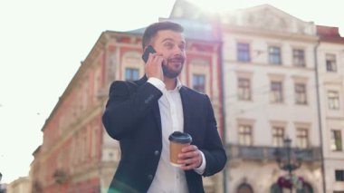 Yakın plan, genç beyaz adamın sinematik portresi. Yürüyor ve telefonda konuşuyor. Kamera 360 çekim. Takım elbiseli adam. Kahve içen bir erkek. Güneş ışığı parlıyor.
