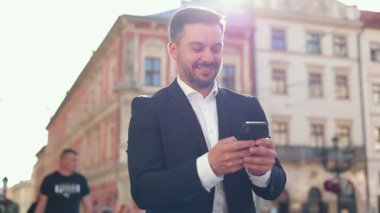 Genç, yakışıklı, beyaz bir adamın yürüdüğü, telefonları kaydırdığı ve binalara baktığının sinematik portresini yakından çek. Kamera 360 çekim. Takım elbiseli adam. Güneş ışığı parlıyor.