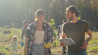 Beyaz bir çift gönüllü çevre gönüllüsü arka planda diğer insanlarla birlikte yürüyor, bazı belgeleri izliyor ya da plan yapıyor ve ağaç dikmeyi tartışıyorlar..