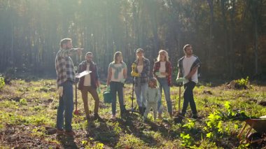 Man curator of eco activists standing with document or plan in hands and giving orders to people for planting trees in garden or forest. Outdoor. Enviroment concept. Working in wood as volunteers.