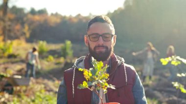 Close up of tree seedling in pot in hands of handsome Caucasian male in glasses, volunteer and gardener. Sunny day outdoor in garden. Portrait of man in eyeglasses with plant. Ecosytem concept.