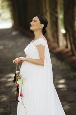 Şaşılacak derecede güzel bir gelin dalgalı beyaz bir elbiseyle süslenmiş, doğanın ortasında saf neşe ve sükunet yayıyor.