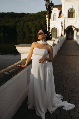 Güzel beyaz bir elbise giymiş, göz kamaştırıcı bir gelin, zarif bir şekilde güzel bir köprünün yanında poz verir.