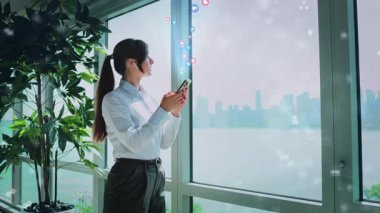 Şehir manzaralı modern bir ofiste, bir kadın akıllı telefonunu kullanır, bağlantı ve kentsel yaşam tarzını gösterir.