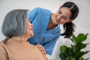 A caregiver rest her hands on the shoulders of a contented senior patient while she sitting on the sofa at home.