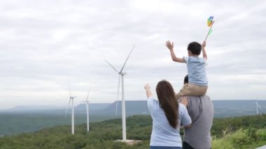 İlerici mutlu aile kavramı rüzgâr türbini çiftliğinde eğleniyor. Rüzgar türbini jeneratörüyle gelen elektrik jeneratörü kırsal bölgede tepe ve ufukta dağ var..