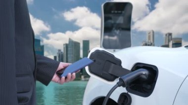 İlerici işadamı elektrikli aracı şarj kablosu ile şarj etme istasyonu ve temiz enerji kampanyası için şehir arka planına sahip akıllı telefondan enerji durumunu kontrol etme.