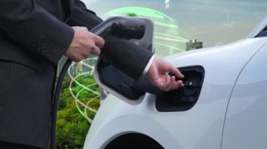 İlerleyen yeşil şehir ESG sembolü, elektrikli araçla birlikte. İş adamı EV arabalarını şarj etmek için şarj aletini şarj ediyor. Şehirdeki şarj istasyonunda ekolojik ve yeşil semboller var..