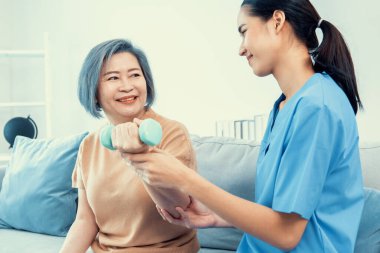 Caregiver helping contented senior woman exercise with dumbbell at home. Rehab for elder, senior medical care.